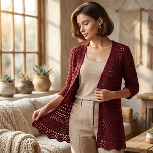 May include: A burgundy open-front crochet cardigan with a scalloped hem. The long-sleeved sweater features an open, airy design. The model is wearing a beige top and pants. The background includes a window, plants, and a sofa.