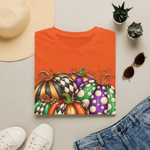 May include: An orange t-shirt featuring a graphic print of four pumpkins with varied patterns including black and white checkered, purple with white polka dots, and orange and green stripes, all topped with brown and green stems.