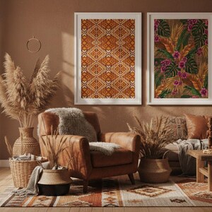 Warm-toned boho living room with two framed African art prints on an earthy terracotta wall. Left: geometric textile pattern in orange and brown. Right: tropical botanical with monstera leaves and pink flowers. Styled with pampas grass and leather armchair
