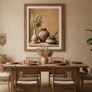 African pottery still life art print framed above a boho dining table. The artwork features traditional clay vases and dried botanicals in warm brown and terracotta tones. Natural wood dining set with woven chairs completes the earthy Afrohemian aesthetic