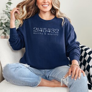 May include: A navy blue crew neck sweatshirt featuring the white text "2145432 worthy & wealthy". The sweatshirt is worn by a person. The person is wearing light blue jeans with ripped knees.