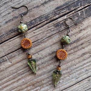 May include: A pair of dangle earrings featuring bronze-toned metal hooks. Each earring consists of a green flower-shaped bead, a round orange bead with a sunburst pattern, a small amber-colored bead, and a translucent green leaf-shaped glass bead.
