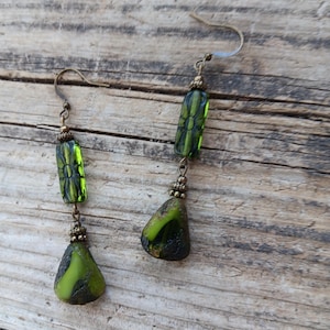 May include: A pair of handcrafted earrings featuring green and black glass beads. The earrings have a hook closure and consist of a rectangular bead, a small decorative element, and a teardrop-shaped bead. The beads have a textured appearance.