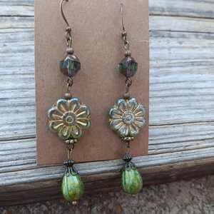 May include: A pair of handcrafted earrings featuring a floral design. The earrings have a bronze-colored hook, a faceted bead, a flower-shaped bead, and a green teardrop-shaped bead. The earrings are displayed on a brown card.