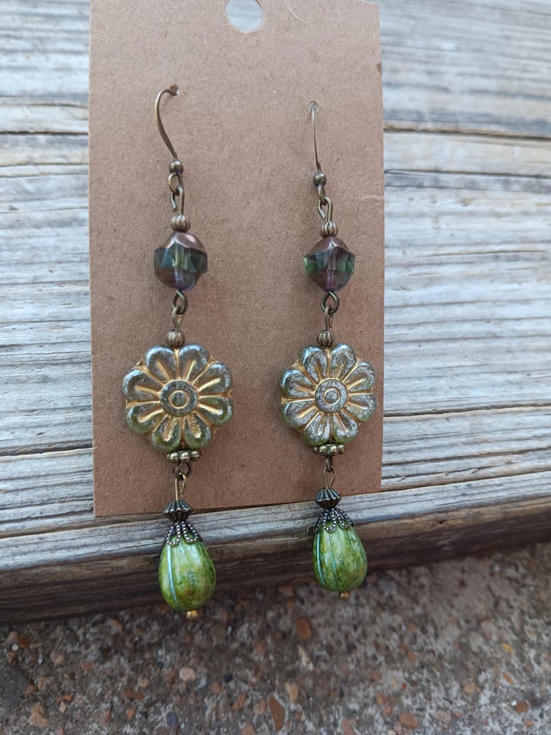 May include: A pair of handcrafted earrings featuring a floral design. The earrings have a bronze-colored hook, a faceted bead, a flower-shaped bead, and a green teardrop-shaped bead. The earrings are displayed on a brown card.