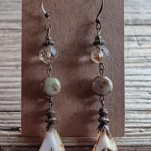 May include: A pair of dangle earrings featuring bronze-toned metal hooks and spacers. Each earring includes a faceted clear bead, a round mottled green and brown stone bead, and a bottom teardrop-shaped bead with white and amber-colored glass.