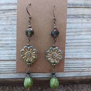 May include: A pair of dangle earrings with a floral theme. The earrings feature a gold-colored flower-shaped bead, green teardrop-shaped beads, and faceted green beads. The earrings are suspended from bronze-colored ear wires.