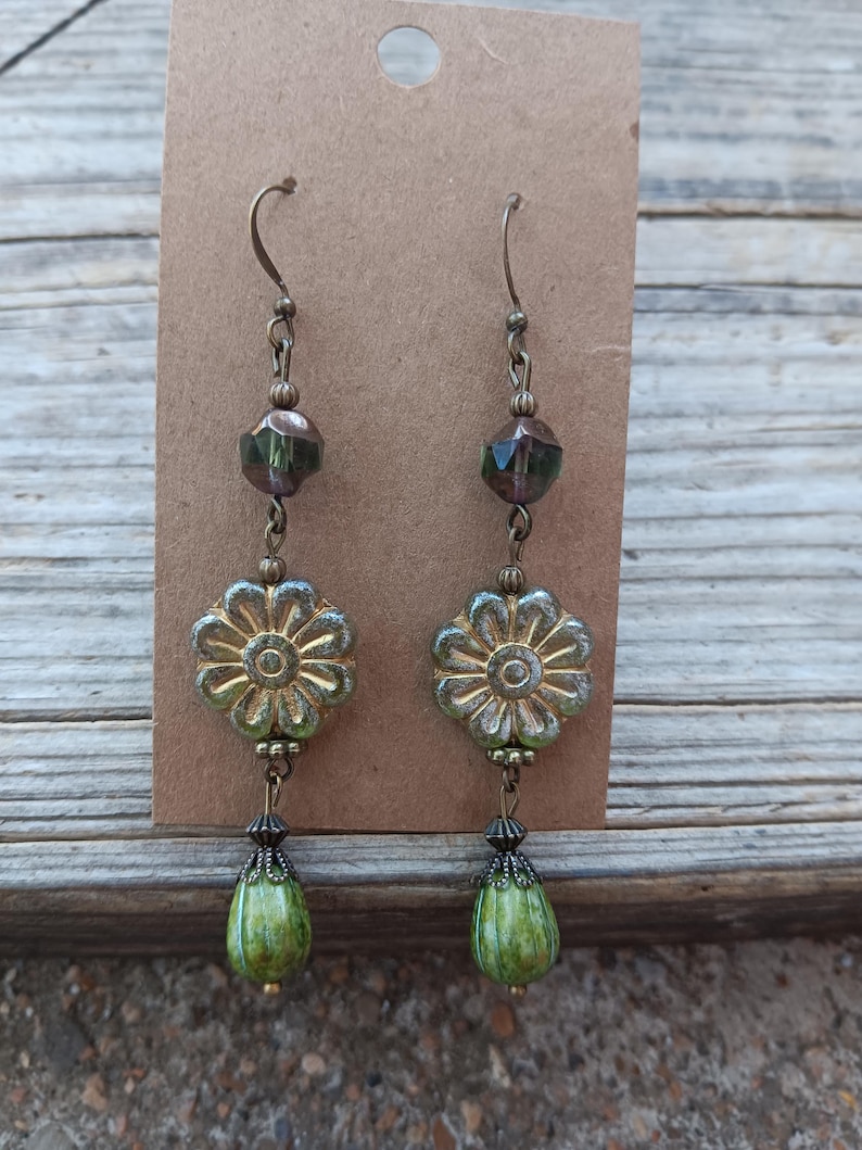 May include: A pair of dangle earrings with a floral theme. The earrings feature a gold-colored flower-shaped bead, green teardrop-shaped beads, and faceted green beads. The earrings are suspended from bronze-colored ear wires.