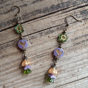May include: A pair of handcrafted earrings with a vintage aesthetic. Each earring features a brass hook, a green flower-shaped bead, a lavender disc with a gold bird design, and a cluster of flower-shaped beads in gold, lavender, and green.