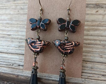 May include: A pair of handcrafted earrings featuring a butterfly and bird design. The earrings have a dark color scheme with black and copper accents. The butterfly and bird charms are suspended from bronze-colored hooks, with dangling black teardrop-shaped beads.
