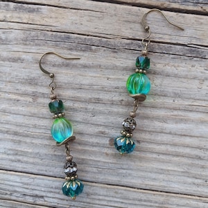 May include: A pair of handcrafted earrings featuring a combination of green and teal beads. The earrings have a vintage brass finish, with a mix of round and faceted beads. The design includes a hook closure and decorative accents, creating a unique accessory.