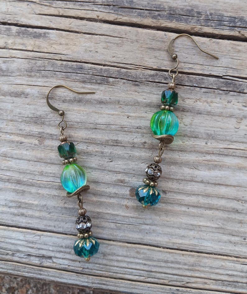 May include: A pair of handcrafted earrings featuring a combination of green and teal beads. The earrings have a vintage brass finish, with a mix of round and faceted beads. The design includes a hook closure and decorative accents, creating a unique accessory.
