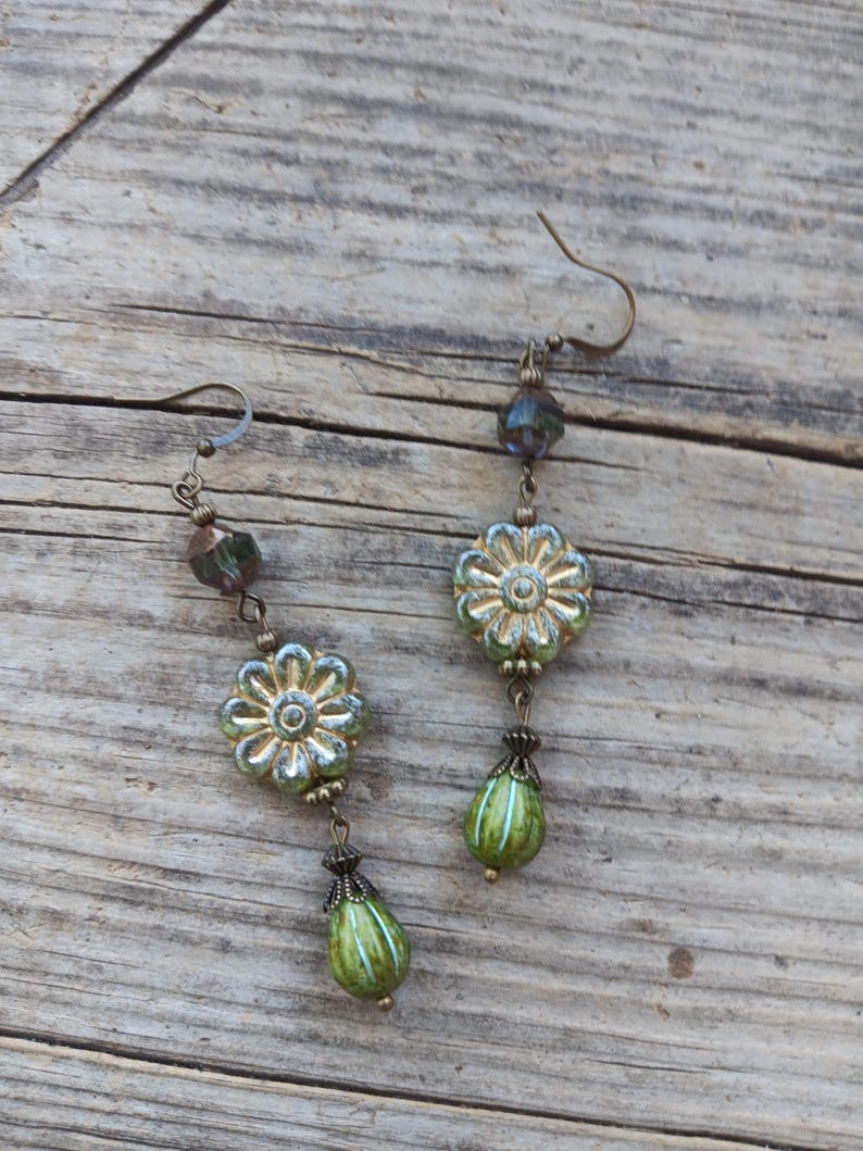 May include: A pair of dangle earrings with a floral design. The earrings feature a gold-colored metal with green and blue glass beads. The earrings have a hook closure and a teardrop-shaped bead at the bottom.