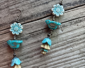 May include: A pair of dangle earrings featuring a white flower bead, a turquoise bird bead with gold accents, and a tiered blue and beige bell-shaped flower bead. The components are connected by small silver-toned metal spacers and hooks.