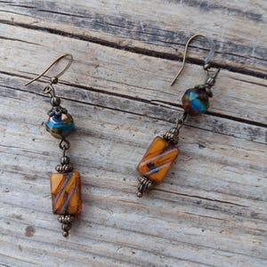 May include: A pair of handcrafted earrings featuring a combination of orange and blue glass beads. The earrings have a vintage aesthetic with bronze-colored metal accents and ear wires. The rectangular and round beads are strung together, creating a unique and artistic design.