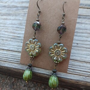 May include: A pair of dangle earrings with a floral theme. The earrings feature a gold-colored flower, a green teardrop-shaped bead, and a faceted bead. The earrings are suspended from a bronze-colored hook.