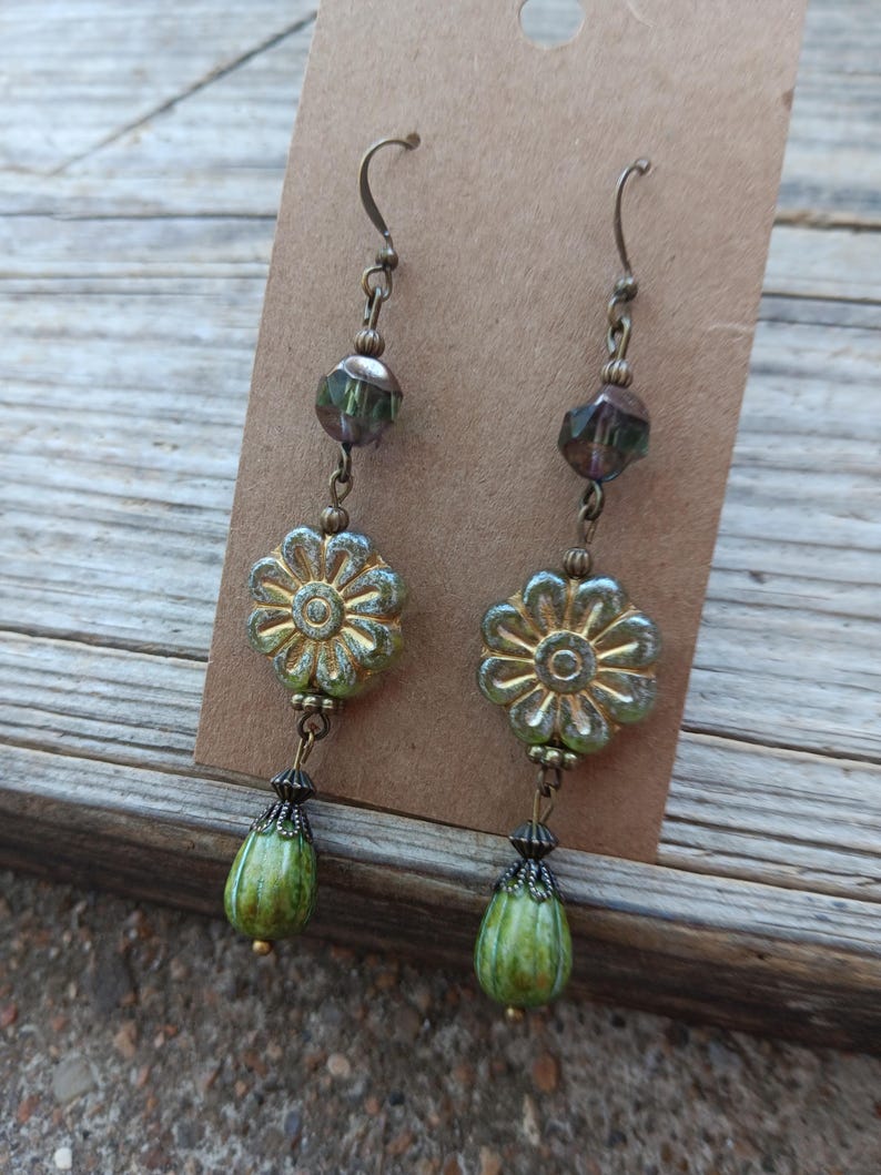 May include: A pair of dangle earrings with a floral theme. The earrings feature a gold-colored flower, a green teardrop-shaped bead, and a faceted bead. The earrings are suspended from a bronze-colored hook.