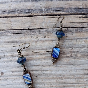 May include: A pair of dangle earrings featuring blue and white striped rectangular beads, small blue round beads, and antique bronze-colored metal accents. The earrings have a hook closure and are set against a rustic wooden background.