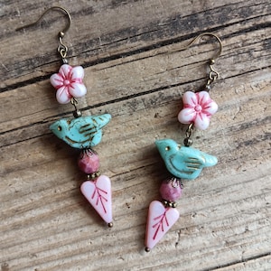 May include: A pair of handcrafted earrings featuring a bird and floral design. The earrings have bronze-colored hooks, with a white and pink flower, a turquoise bird, a pink bead, and a pink heart-shaped charm. The earrings are on a wooden surface.