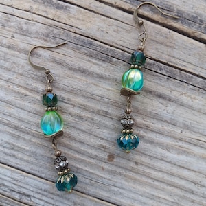 May include: A pair of dangle earrings with an antique brass finish. Each earring features a combination of teal and green glass beads, with a faceted teal bead at the bottom and a decorative bead with rhinestones.