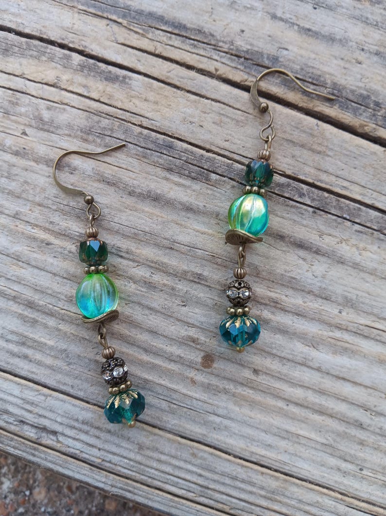 May include: A pair of dangle earrings with an antique brass finish. Each earring features a combination of teal and green glass beads, with a faceted teal bead at the bottom and a decorative bead with rhinestones.