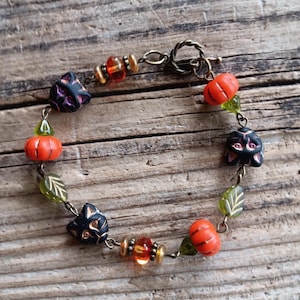 May include: A bracelet featuring black cat-shaped beads, orange pumpkin-shaped beads, translucent green leaf-shaped beads, and small amber-colored glass beads with metallic spacers, all connected by a bronze-toned metal chain with a toggle clasp.