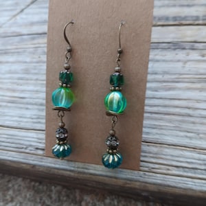 May include: A pair of dangle earrings with a vintage aesthetic. Each earring features a bronze-colored hook, green and teal beads, and decorative accents. The earrings are displayed on a brown card against a wooden background.