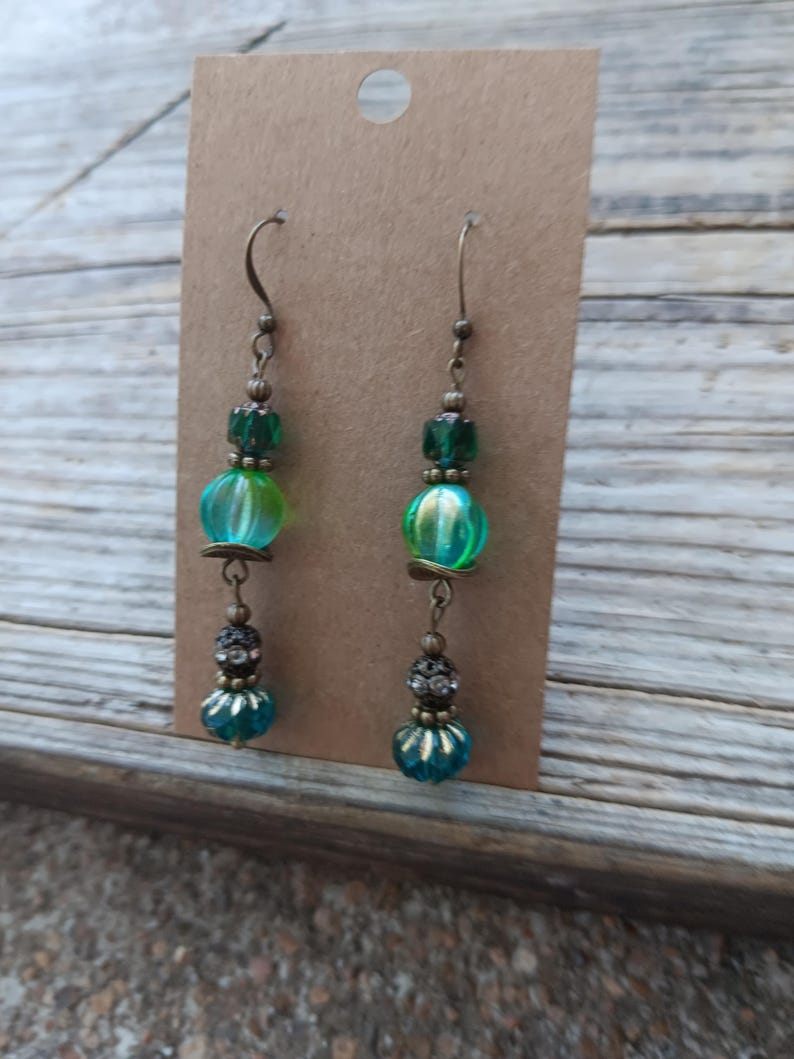 May include: A pair of dangle earrings with a vintage aesthetic. Each earring features a bronze-colored hook, green and teal beads, and decorative accents. The earrings are displayed on a brown card against a wooden background.