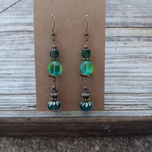 May include: A pair of handcrafted earrings with a vintage aesthetic. Each earring features a combination of green and teal beads, including a large, translucent green bead. The earrings are suspended from bronze-colored hooks and are displayed on a brown card.
