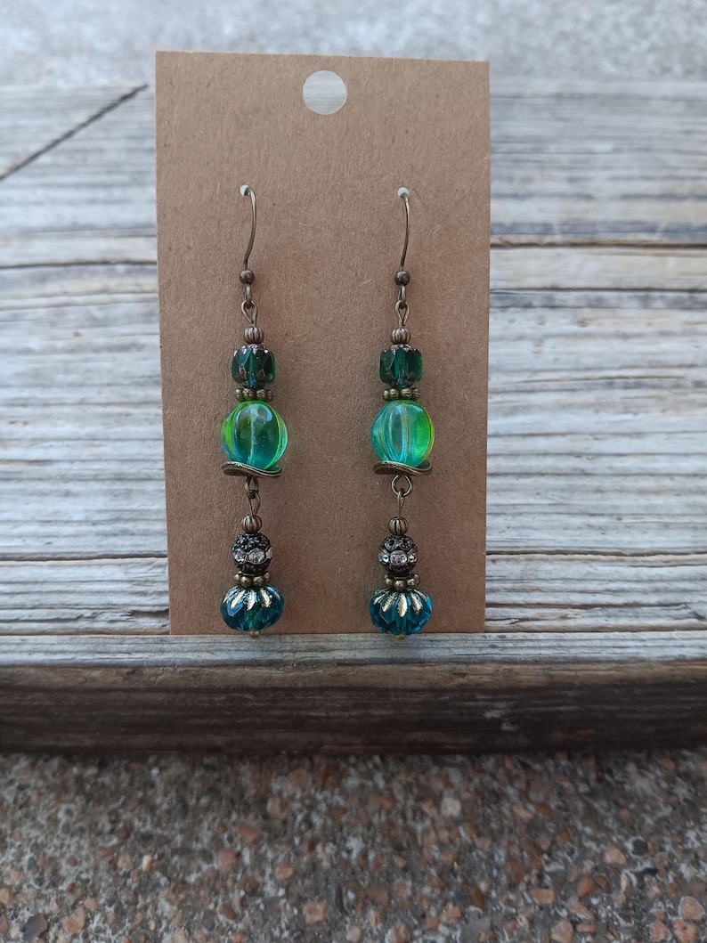 May include: A pair of handcrafted earrings with a vintage aesthetic. Each earring features a combination of green and teal beads, including a large, translucent green bead. The earrings are suspended from bronze-colored hooks and are displayed on a brown card.