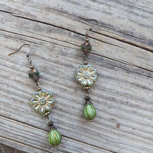 May include: A pair of dangle earrings featuring a floral design. The earrings have a silver-tone metal hook and are adorned with light green flower-shaped beads, green teardrop beads, and smaller green and brown beads. The earrings are displayed on a wooden surface.