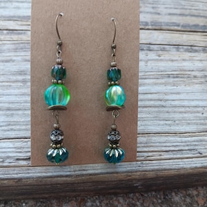 May include: A pair of dangle earrings with a vintage aesthetic. Each earring features a combination of teal, green, and clear beads, with antique bronze-colored metal accents. The earrings are displayed on a brown card against a wooden background.