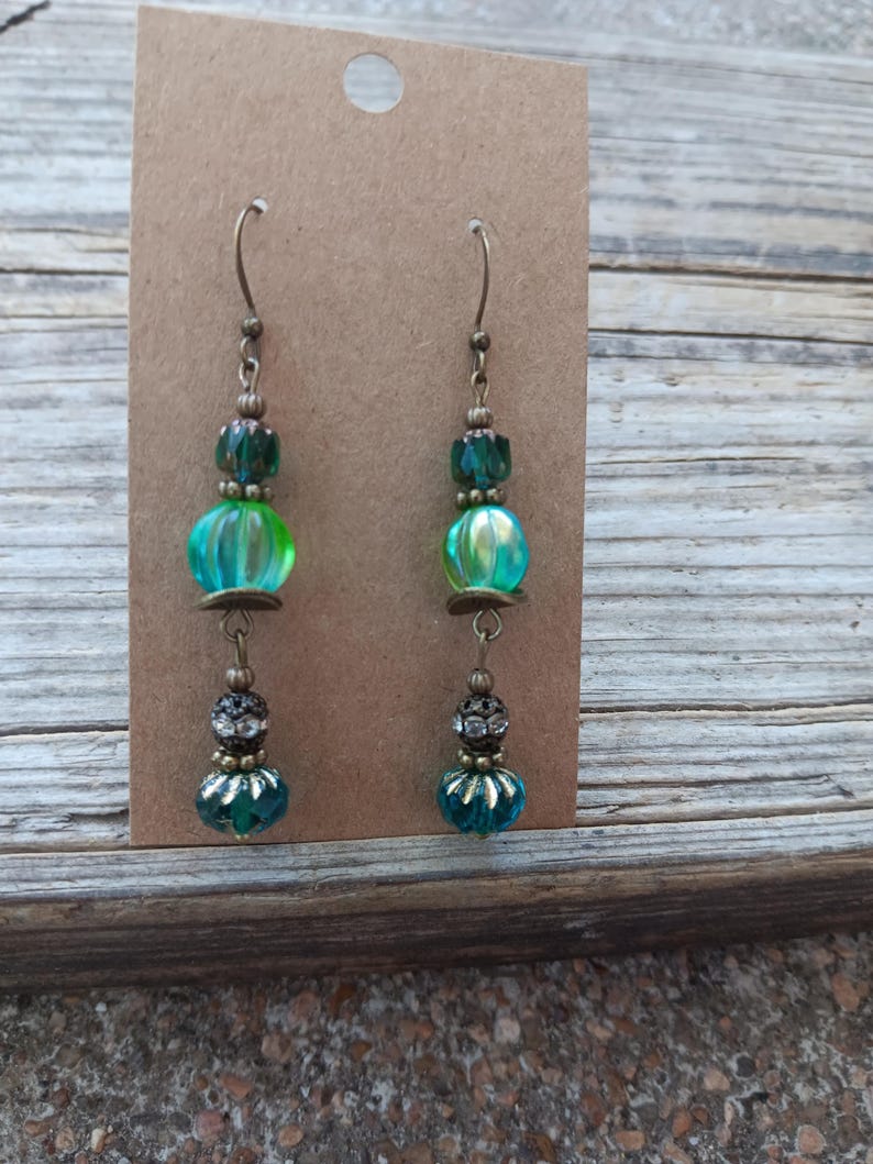 May include: A pair of dangle earrings with a vintage aesthetic. Each earring features a combination of teal, green, and clear beads, with antique bronze-colored metal accents. The earrings are displayed on a brown card against a wooden background.