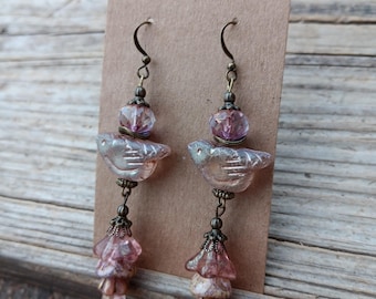 May include: A pair of handcrafted earrings featuring a bird design. The earrings have a clear, frosted bird-shaped bead, a purple bead, and a pink flower-shaped bead. The metal components have an antique bronze finish.