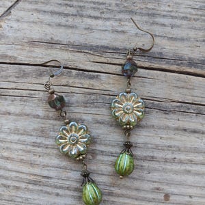 May include: A pair of handcrafted earrings featuring a floral design. The earrings have a bronze-colored hook and are adorned with green and gold-colored flower-shaped beads, along with green teardrop-shaped beads. The earrings are displayed on a wooden surface.