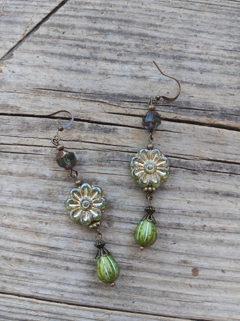 May include: A pair of handcrafted earrings featuring a floral design. The earrings have a bronze-colored hook and are adorned with green and gold-colored flower-shaped beads, along with green teardrop-shaped beads. The earrings are displayed on a wooden surface.