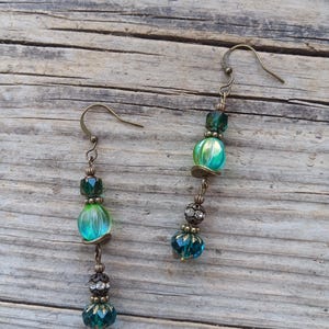 May include: A pair of dangle earrings with a vintage aesthetic. Each earring features a fish hook, connected to a series of beads in shades of green and teal. The beads include faceted, round, and decorative elements, with a bronze-colored metal finish.