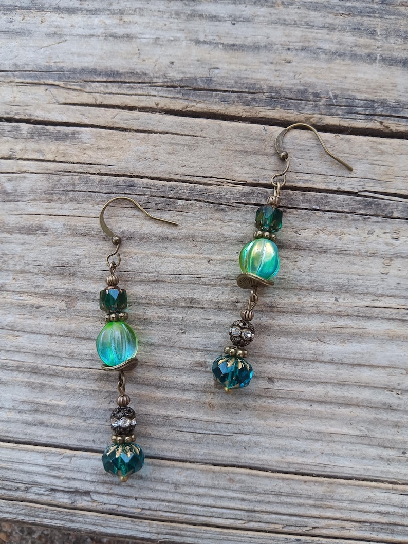 May include: A pair of dangle earrings with a vintage aesthetic. Each earring features a fish hook, connected to a series of beads in shades of green and teal. The beads include faceted, round, and decorative elements, with a bronze-colored metal finish.
