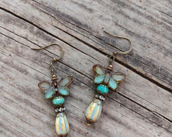 May include: A pair of butterfly earrings with a vintage aesthetic. The earrings feature turquoise and cream-colored beads, with a butterfly design in shades of blue and brown. The earrings are suspended from gold-colored hooks.