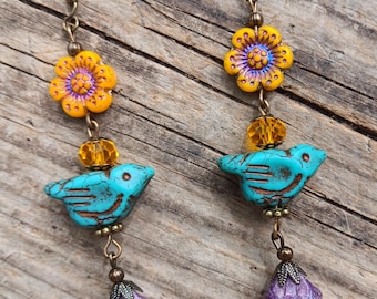 May include: A pair of handcrafted earrings featuring a floral design with yellow and purple accents. Each earring has a turquoise bird charm, a cluster of colorful beads, and a purple flower charm. The earrings are suspended from bronze-colored hooks.