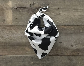 May include: A white and black cow print bandana, knotted at the top. The bandana is made of a soft fabric and is hanging against a weathered wood background. The bandana is a triangular shape.