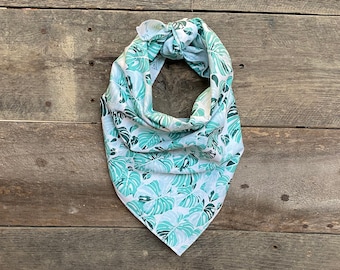 May include: A white and teal bandana with a tropical leaf pattern. The bandana is knotted at the top and triangular in shape. The leaves are in shades of teal and green, set against a white background. The bandana is made of soft fabric.