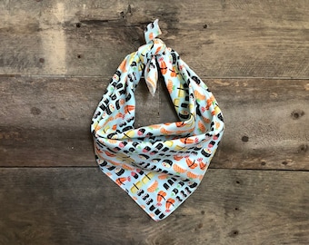 May include: A light blue dog bandana with a repeating pattern of sushi rolls in black, orange, and yellow. The bandana is knotted at the top and is made of a soft fabric. The background is a rustic wooden surface.