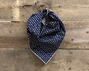 May include: A navy blue bandana with a white arrow pattern. The bandana is knotted and has a white border. The fabric is folded and hangs against a wooden background.