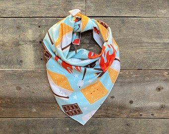 May include: A light blue bandana with a repeating pattern of s'mores, campfires, and stars. The bandana is tied at the top, showcasing the colorful design. The design includes marshmallows, chocolate bars, and graham crackers.