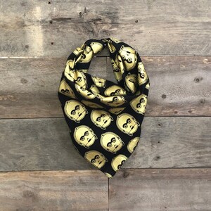 May include: Black and gold patterned bandana featuring a repeating design of a robot's face. The bandana is folded and displayed against a weathered wood background. The fabric has a shiny appearance.