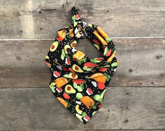 May include: Black dog bandana with a colorful taco pattern. The bandana features illustrations of tacos, avocados, tomatoes, and limes. The fabric is knotted at the top, creating a triangular shape.