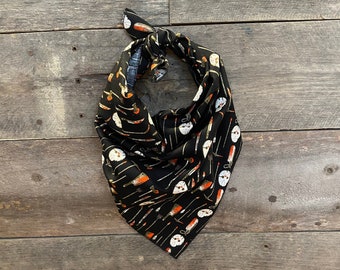 May include: Black bandana with a repeating pattern of white hockey masks, cleavers, and knives. The bandana is tied at the top and is made of a soft fabric. The background is a rustic wooden surface.