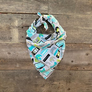 May include: A gray bandana with a pattern of video game controllers, consoles, and screens in shades of blue, green, and black. The bandana is tied at the top and hangs in a triangular shape.
