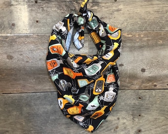 May include: Black Halloween-themed dog bandana with a repeating pattern of potion bottles, spiderwebs, and text. The bottles are in various colors, including orange, green, and white. The bandana is tied at the top.